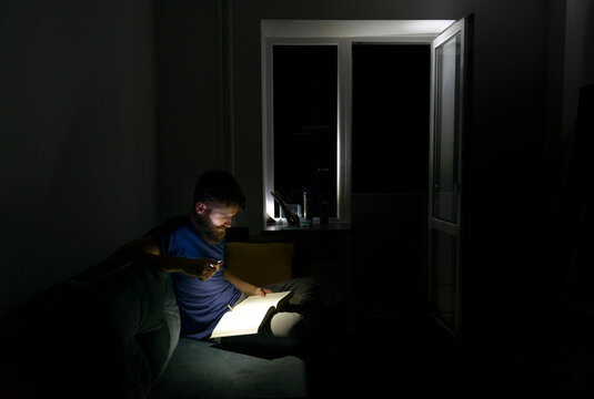 Man reading book with flashlight during power outage at night, sitting on sofa in dark room