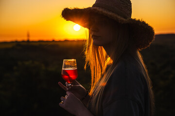 Teenage girl wearing a straw hat holds a glass of red drink and looks at the sunset in a peaceful...