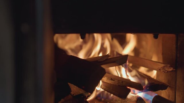 Extreme close up of individual using blue gas torch to ignite stacked firewood inside dark fireplace, flames grow gradually, illuminating wooden logs with warm flickering firelight and glowing embers