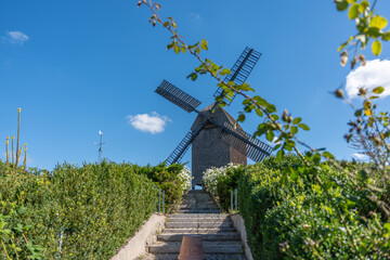An old windmill under a blue sky, surrounded by green hedges and flowers. A staircase leads...