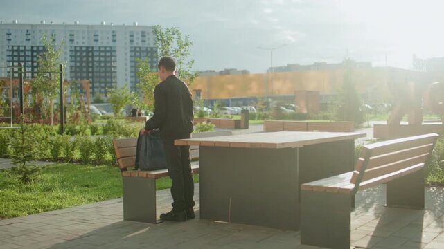 Student standing by bench outdoors zipping backpack after finishing study, preparing to walk away in bright daylight with green plants and modern buildings visible in background