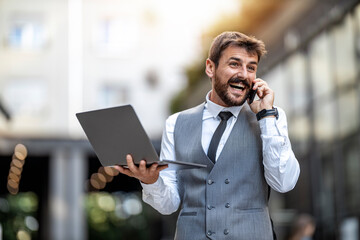 Businessman in a suit talking on the phone while holding a laptop outdoors in a city setting during...