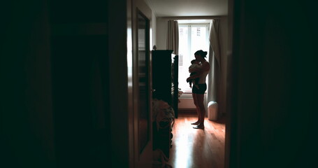 Wide shot of mother standing in room, holding baby while natural light from window illuminates the...