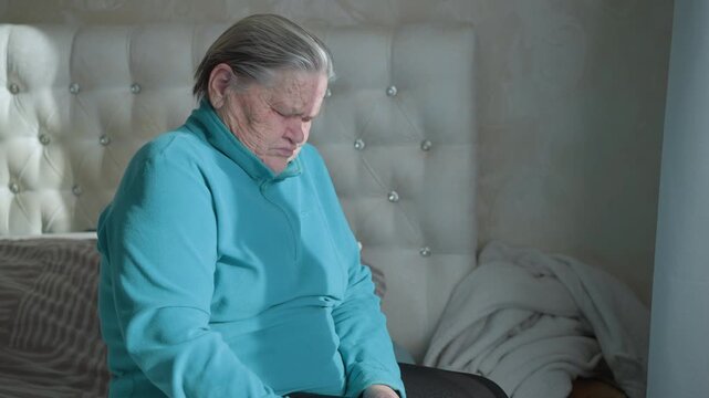 Old woman in green sweatshirt sitting on bed, holding her lower back, wincing in pain. She s experiencing discomfort, possibly from back pain, in her home while dressed casually in comfortable