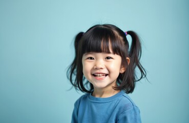 Cheerful baby Asian toddler girl with adorable bangs, pigtails wears blue sweater, smiling joyfully at camera. Cute child represents innocence, happiness, childhood. Simple studio background enhances