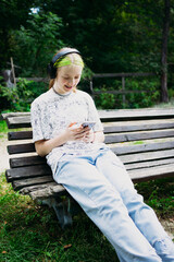 Teen girl with bright green hair smiling while looking at smartphone and listening to music in black headphones. Sitting on a bench in the park, enjoying leisure and modern lifestyle