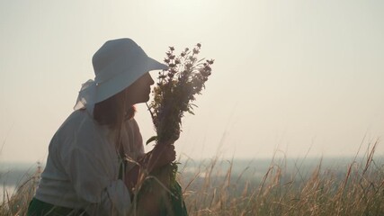 woman in white sunhat squatting in tall grass holding bouquet of wildflowers close to face while gently inhaling scent under soft sunlight with serene sky and golden field background