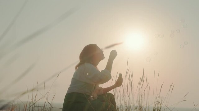 Silhouetted woman crouches in tall grass blowing bubbles toward glowing sunset sky, warm light casting long shadows across field, floating bubbles catching soft hues