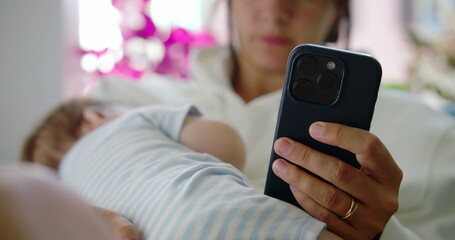 Mother holding her baby on her lap while browsing her phone, with the baby peacefully resting in her arms in a relaxed, brightly lit home