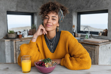 Smiling woman in mustard sweater sitting in rustic kitchen with smoothie bowl and orange juice, modern healthy lifestyle and wellness concept with natural light and cozy atmosphere