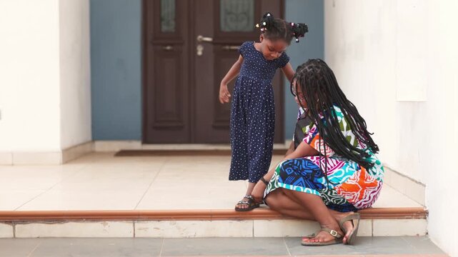 Family moment, African mother fastens her daughter's sandal at the front of their home.