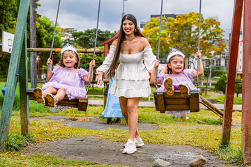 Mother plays with twin daughters on swings during outdoor birthday celebration in park.