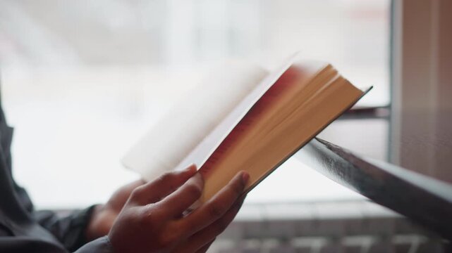 Close-up of hands holding open book in front of window during snowy day, pages turning mid-motion, soft natural light, calm indoor scene with student reading quietly, peaceful focus on printed text