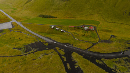 Aerial image of a rural Icelandic area featuring a red roofed house, green fields, volcanic soil, a winding road, vehicles, and rolling hills.