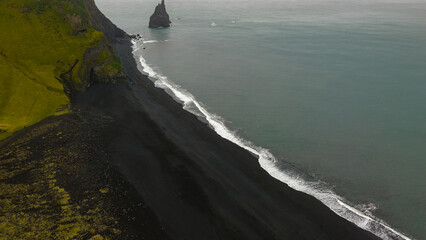 Aerial view of a black sand beach with green cliffs, a sea stack near the shore, and the ocean stretching into the distance along Iceland's coastline.