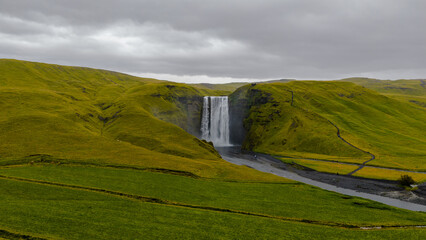 Fototapeta premium Skogafoss waterfall flows powerfully from a cliff, surrounded by green hills and a misty spray. A winding path and dark riverbed complete the dramatic scene.