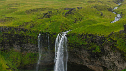 Seljalandsfoss waterfall cascades from a tall cliff, surrounded by green moss and grass, with a stream winding through the lush Icelandic terrain. © Ekaterina
