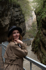 A stylish woman in a beige trench coat and black hat leans on a railing in a mountain canyon, smiling softly. Adventure, female travel, exploration