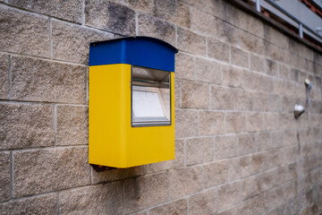 Yellow and blue public mailbox attached to brick wall, representing postal system process, communication infrastructure, mail delivery equipment, and traditional correspondence service