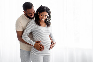 Fototapeta premium Satisfied millennial black handsome hugs pregnant wife in bedroom interior and look at belly on window background. Couple awaiting baby, cute parents-to-be together at home, family enjoy tender moment