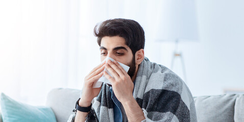 Ill arab man covered in warm blanket looking at laptop screen and sneezing nose in paper tissue, sitting on sofa at home, empty space. Sinusitis illness, cold and influenza symptoms