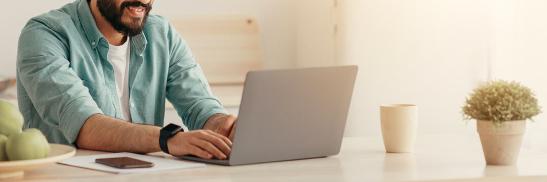 Arab guy spending time at home, using laptop computer, sitting in kitchen interior, copy space. Happy man surfing on Internet, typing on computer keyboard, chatting online - Powered by Adobe