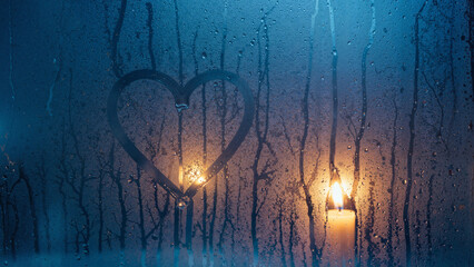 Heart shape drawn on foggy glass with candles glowing in the dark  
