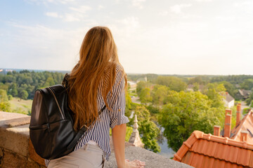 Young woman with backpack enjoys panoramic view over rooftops, forest and horizon, capturing the...
