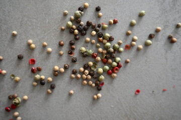 A top-down shot of spilled mixed colourful peppercorns, including black, white, green, and red varieties, on a grey textured surface.