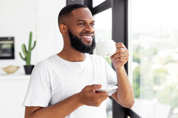 Good Morning. Portrait Of Smiling Relaxed African American Man Enjoying Start Of The Day, Drinking Hot Coffee Standing And Looking Through The Window At Home, Holding Plate And Cup With Beverage