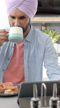 Vertical video: Man is browsing tablet and sipping coffee with toast on counter during breakfast