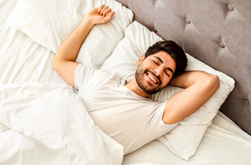 Good morning. Happy wellslept arab guy waking up in the morning and stretching hands, lying in...