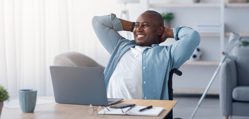 Obraz premium Rest and relax at home, work remotely, successful business and break. Happy mature african american guy disabled in wheelchair threw hands behind head at table with laptop in living room interior