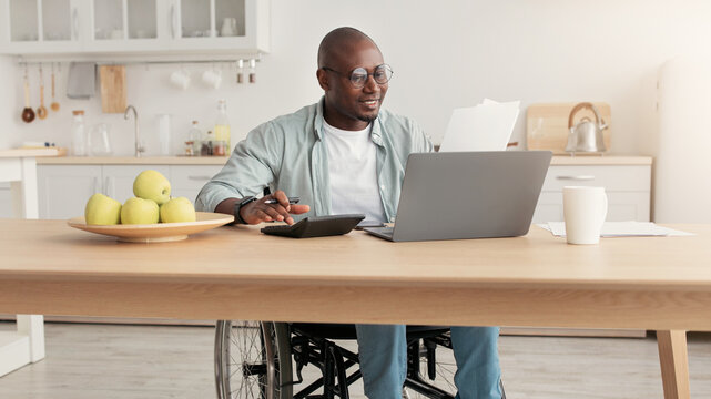 Paperwork, payment of bills, taxes, home bookkeeping and manager work. Cheerful middle aged african american man disabled in glasses in wheelchair counts money on calculator and laptop indoors