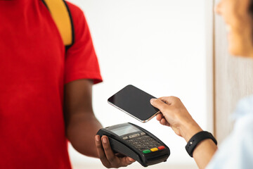 Closeup of woman using mobile phone with blank screen for contactless payment, black male courier thermo backpack holding Point Of Sale machine and cardboard box with purchase order, selective focus