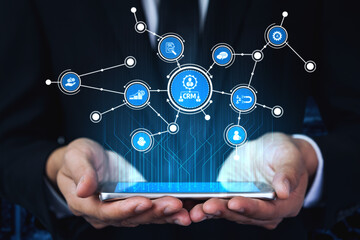 A businessman holds a smartphone displaying icons representing CRM and digital connectivity in a modern office environment, symbolizing innovation in business management. Tessel