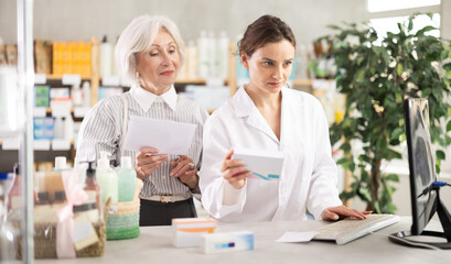 Woman employee in labcoat works in pharmacy using computer and collaborate with senior female client. Professional comment doctors prescription, recommend drug for treatment regiment