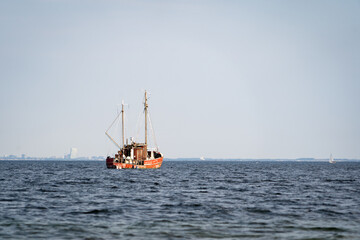 Red fishing boat with twin masts on calm blue sea, distant city skyline on the horizon, daytime seascape © Piotr