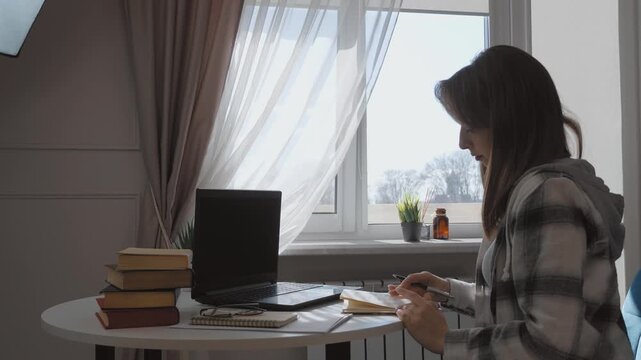 Young female student diligently taking notes from laptop screen, writing in notebook while preparing for exams and studying at home with focused concentration
