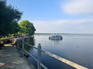 Naklejka premium A serene lakeside view with a boat on calm water. Trees line the shore, and a walkway runs alongside the lake, providing a peaceful atmosphere.