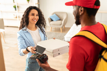 Fast Food Delivery, Digital Payment. Smiling woman using mobile phone, black male courier in red uniform holding Point Of Sale atm terminal in hand and giving pizza boxes to happy consumer