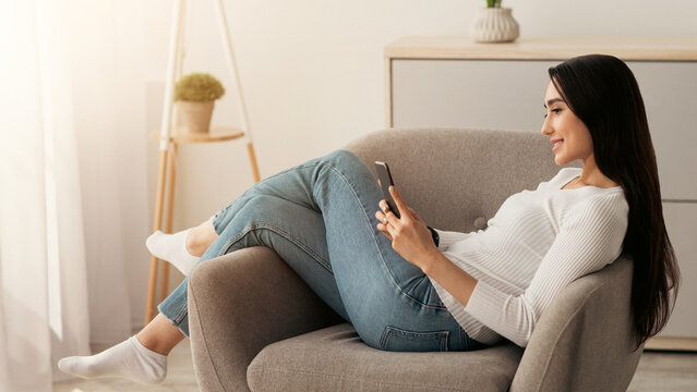 Rest at home, social networks, modern chat, surfing in online, enjoy free time. Smiling young pretty arab woman reading message on smartphone in armchair, in minimalist living room interior, profile
