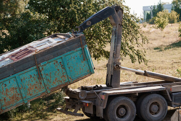 Rusty Dump Truck Lifting Waste Container with Crane Arm in Rural Field Landscape, Sunlit Vintage...