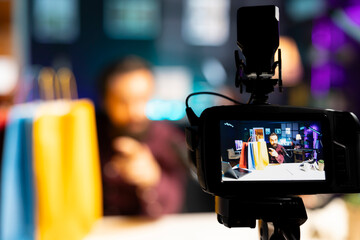 Close up shot of professional camera used by media star holding colorful shopping bags in hand to filming vlog. Focus on specialized gear used by content creator in blurry background