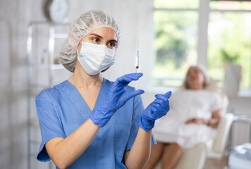Female doctor holding syringe in medical mask and gloves, for injecting the vaccine into patient who is sitting behind.