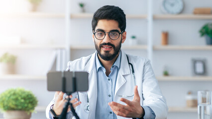 Medical blog, webinar and online consultation during covid-19 social distancing. Friendly 30s arabian doctor in coat and glasses, gesturing and looking at phone webcam at table in office interior