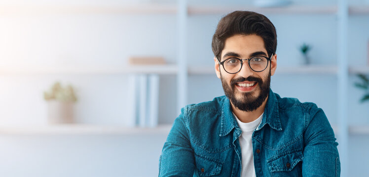 Happy toung arab self-entrepreneur man sitting at desk in home office interior and smiling to camera. Handsome millennial businessman posing at workplace, copy space