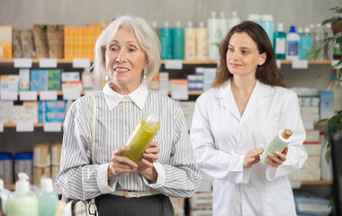 Senior woman chooses a medicated shampoo for gray and damaged hair in a European pharmacy. Female pharmacist helps a pensioner with the choice of medical shampoo for hair treatment
