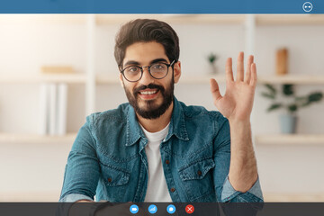 Hello. Happy arab man waving hand and smiling to camera, sitting at workplace at home. Eastern guy gesturing hi and greeting while making video call. Modern remote communication concept © Prostock-studio