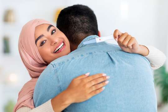 Excited muslim woman with positive pregnancy test hugging her husband, smiling at camera, living room interior. Young arab family enjoying parenthood, embracing at home - Powered by Adobe